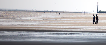 Sun sea sand shadow 2 This landscape photograph depicts Sutton on Sea Beach in Lincolnshire, England, on a late spring morning. The main subject, titled "Sun sea sand shadow 2," shows people walking along the expansive sandy beach as the sunlight creates shadow patterns across the sand and shallow tidal pools. The gentle waves of the North Sea can be seen in the background, while distant figures stroll along the shore, illustrating the quiet coastal charm of this United Kingdom location. A prominent beach marker is visible near the horizon, adding a distinctive feature to the seafront scene and enhancing the local identity of Sutton on Sea.
